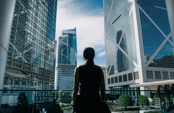 Rear view of professional young businesswoman standing against contemporary financial skyscrapers in downtown financial district and looking up into sky with positive emotion