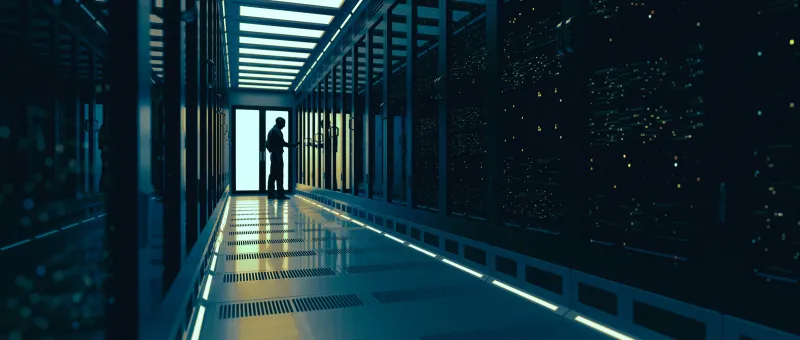 Silhouette of a person walking through a brightly lit server room corridor with tall racks.