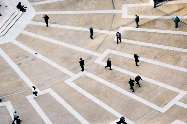 People walking across a large, patterned paved plaza with white lines and tiles.